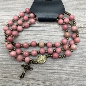 May include: A pink and gold rosary bracelet with a small cross charm and a medal of the Virgin Mary. The bracelet is made of pink stone beads and gold metal beads.