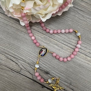 Op de afbeelding: Een roze en witte rozenkrans met een gouden kruis en een gouden medaillon met een religieus beeld. De rozenkrans ligt op een houten oppervlak.