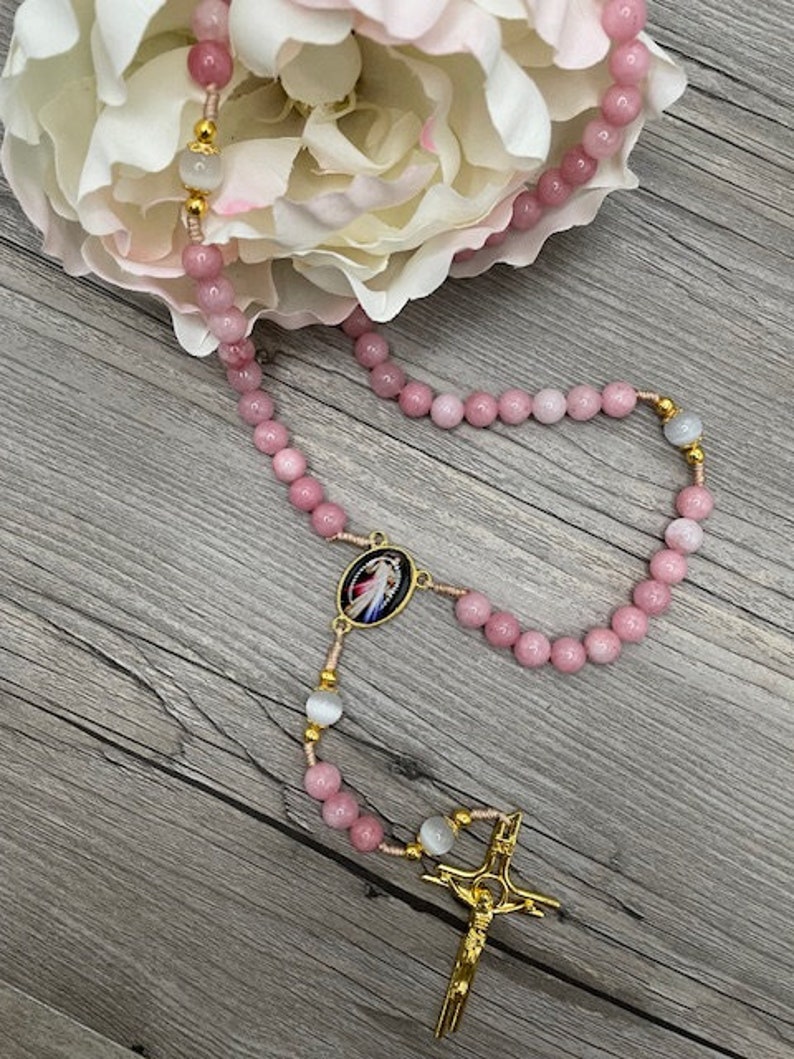 Op de afbeelding: Een roze en witte rozenkrans met een gouden kruis en een gouden medaillon met een religieus beeld. De rozenkrans ligt op een houten oppervlak.