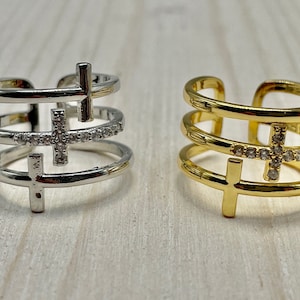 May include: Two adjustable rings with a triple band design. One ring is silver with clear stones and the other is gold with clear stones. Both rings feature a cross design.