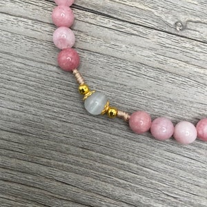 Op de afbeelding: Een roze en witte edelsteen kralenketting met gouden accenten. De ketting is gemaakt met ronde kralen en heeft een enkele grotere witte kraal met gouden accenten.