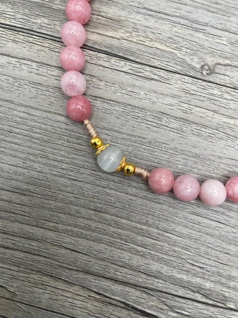 Op de afbeelding: Een roze en witte edelsteen kralenketting met gouden accenten. De ketting is gemaakt met ronde kralen en heeft een enkele grotere witte kraal met gouden accenten.