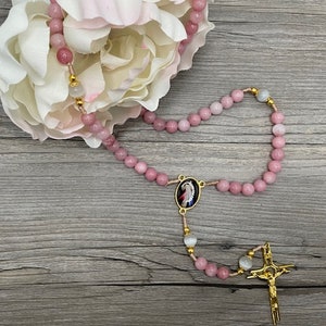 Op de afbeelding: Een roze en witte rozenkrans met een gouden kruis en een klein beeld van de Maagd Maria. De rozenkrans is gemaakt van kralen en is geregen aan een gouden koord.