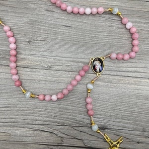 Op de afbeelding: Een roze en witte rozenkrans met een gouden kruis en een ronde hanger met een religieus beeld. De rozenkrans is gemaakt van kralen en is geregen op een dun koord.