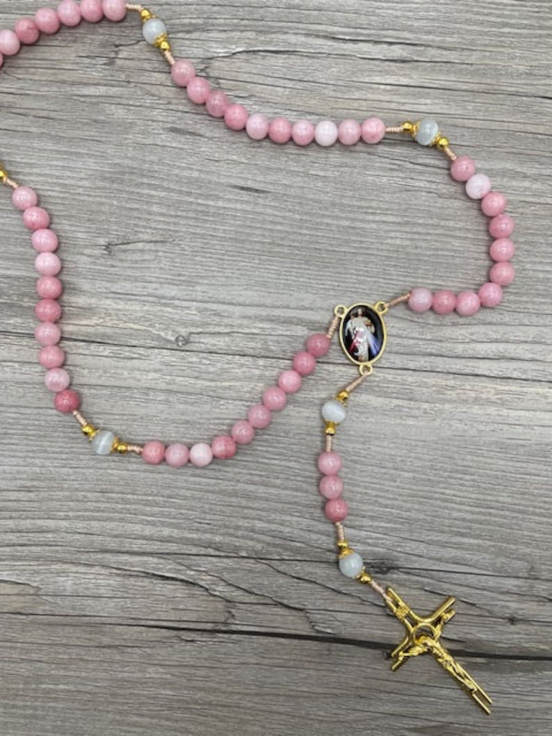 Op de afbeelding: Een roze en witte rozenkrans met een gouden kruis en een ronde hanger met een religieus beeld. De rozenkrans is gemaakt van kralen en is geregen op een dun koord.