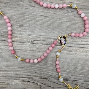 Op de afbeelding: Een roze en witte rozenkrans met een gouden kruis en een gouden ovale hanger met een religieus beeld. De rozenkrans is gemaakt van kralen en is geregen aan een gouden ketting.