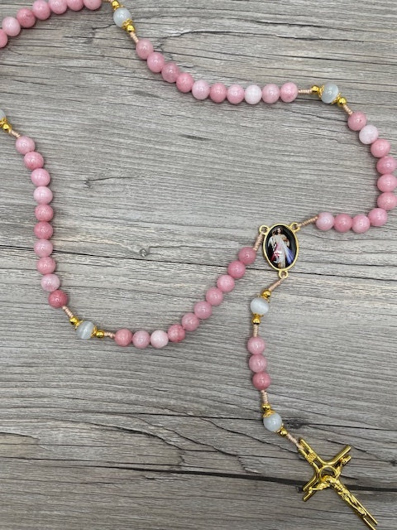 Op de afbeelding: Een roze en witte rozenkrans met een gouden kruis en een gouden ovale hanger met een religieus beeld. De rozenkrans is gemaakt van kralen en is geregen aan een gouden ketting.