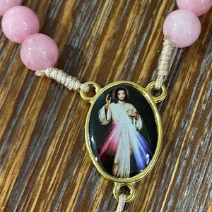 Op de afbeelding: Een roze en witte rozenkrans met een gouden ovale hanger met een afbeelding van Jezus Christus. De hanger heeft een zwarte achtergrond en de afbeelding van Jezus is omringd door een regenboog.