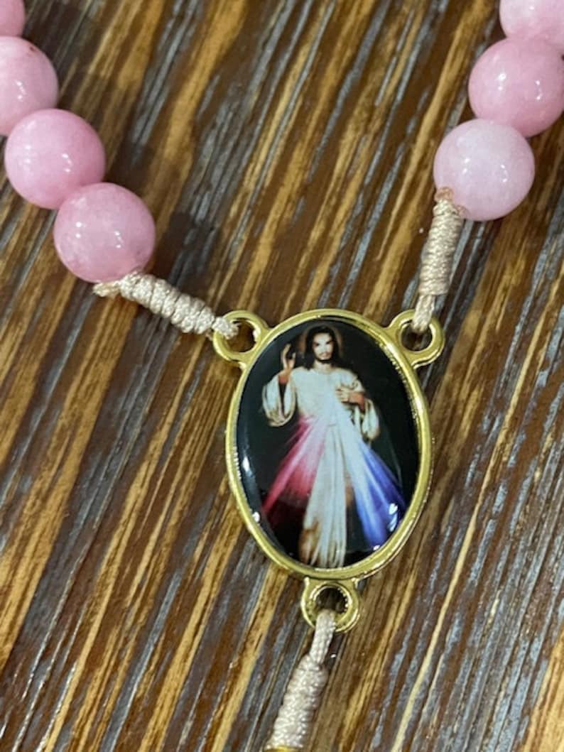 Op de afbeelding: Een roze en witte rozenkrans met een gouden ovale hanger met een afbeelding van Jezus Christus. De hanger heeft een zwarte achtergrond en de afbeelding van Jezus is omringd door een regenboog.