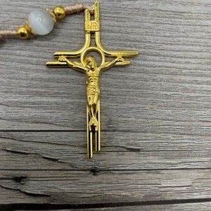 Op de afbeelding: Een gouden metalen kruisbeeld met een figuur van Jezus Christus op het kruis. Het kruisbeeld heeft een eenvoudig ontwerp met een ronde halo rond het hoofd van Jezus. Het kruisbeeld hangt aan een bruin koord.