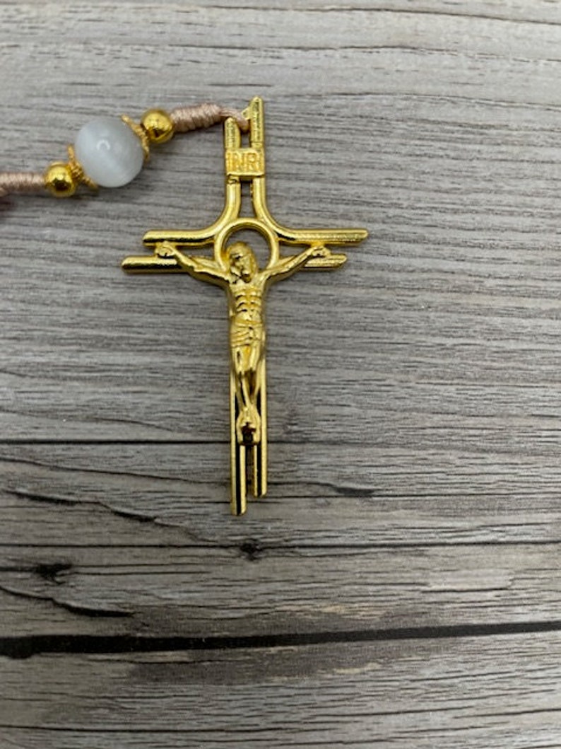 Op de afbeelding: Een gouden metalen kruisbeeld met een figuur van Jezus Christus op het kruis. Het kruisbeeld heeft een eenvoudig ontwerp met een ronde halo rond het hoofd van Jezus. Het kruisbeeld hangt aan een bruin koord.