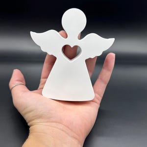 May include: A white angel figurine with a heart-shaped cutout in the center. The angel has outstretched wings and a simple, elegant design. The figurine is held in a hand against a dark background.