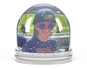 Puede incluir: Un globo de nieve con una foto de un niño que lleva una camisa azul, un sombrero negro y gafas de sol naranjas. El niño sonríe y mira a la cámara. El globo de nieve está lleno de purpurina blanca.