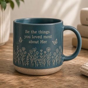 May include: A teal ceramic mug featuring a light-colored speckled base and a handle on the right side. The front displays the text "Be the things you loved most about Her" in a serif font, surrounded by line-art illustrations of wildflowers and butterflies.