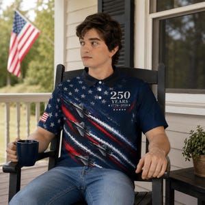 Puede incluir: Polo azul marino con un dise&ntilde;o patri&oacute;tico con estrellas, aviones a reacci&oacute;n y rayas rojas y blancas. La camisa tiene el texto "250 YEARS 1776-2026". Una bandera americana est&aacute; en el fondo.