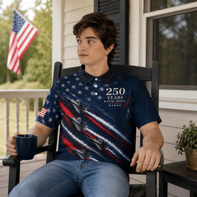 Puede incluir: Polo azul marino con un dise&ntilde;o patri&oacute;tico con estrellas, aviones a reacci&oacute;n y rayas rojas y blancas. La camisa tiene el texto "250 YEARS 1776-2026". Una bandera americana est&aacute; en el fondo.