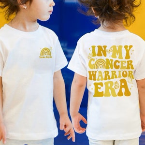 Pu&ograve; includere: Due magliette bianche con testo e grafica dorati. Una maglietta dice "Team Annie" con una grafica arcobaleno. L'altra maglietta dice "In My Cancer Warrior Era" con una grafica arcobaleno, farfalla e nastro.