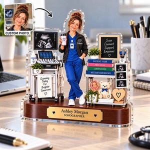 May include: A custom acrylic display featuring a sonographer figurine in blue scrubs and a black jacket, standing beside an ultrasound machine. The base includes a gold plaque reading Ashley Morgan Sonographer, with miniature books, a teddy bear, and ultrasound images.
