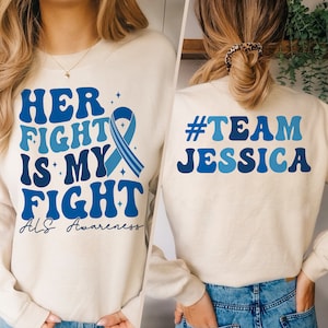 May include: A cream colored sweatshirt with the text "HER FIGHT IS MY FIGHT ALS Awareness" and a blue ribbon on the front. The back of the sweatshirt has the text "#TEAM JESSICA" in blue.
