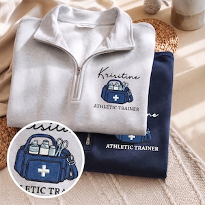 May include: A light gray quarter-zip sweatshirt and a dark navy quarter-zip sweatshirt, each featuring an embroidered blue medical bag graphic with the name Krisitine and the text ATHLETIC TRAINER printed in black lettering on the front chest.