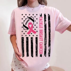 Puede incluir: Camiseta rosa claro con un dise&ntilde;o de concienciaci&oacute;n sobre el c&aacute;ncer de mama. El dise&ntilde;o incluye un bal&oacute;n de f&uacute;tbol, una cinta rosa, estrellas y rayas negras. El texto "TACKLE BREAST CANCER" est&aacute; impreso verticalmente.