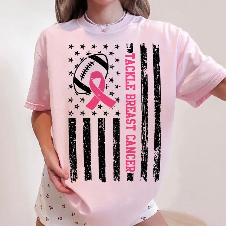 Puede incluir: Camiseta rosa claro con un dise&ntilde;o de concienciaci&oacute;n sobre el c&aacute;ncer de mama. El dise&ntilde;o incluye un bal&oacute;n de f&uacute;tbol, una cinta rosa, estrellas y rayas negras. El texto "TACKLE BREAST CANCER" est&aacute; impreso verticalmente.