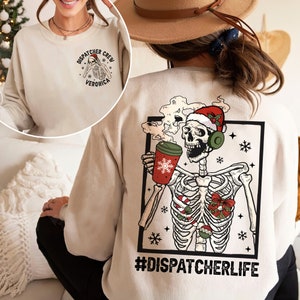 May include: A beige sweatshirt with a Christmas-themed skeleton graphic on the back. The skeleton is wearing a Santa hat and headphones and holding a cup of coffee. The text "#DISPATCHERLIFE" is printed below the graphic. The front of the sweatshirt has a small circle graphic with the text "DISPATCHER CREW VERONICA".