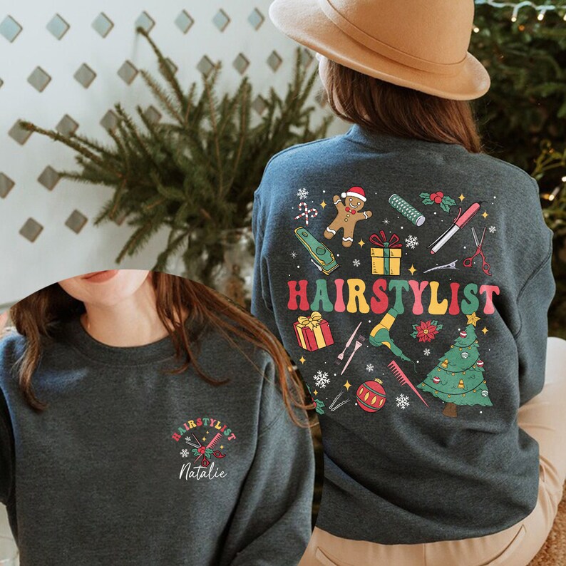 Op de afbeelding: Een donkerblauw sweatshirt met een kerstthema ontwerp met een peperkoekman, een kerstboom en verschillende haarstylingtools. De tekst "HAIRSTYLIST" is in regenboogkleuren gedrukt op de achterkant van het sweatshirt. De voorkant van het sweatshirt heeft een kleiner ontwerp met de tekst "HAIRSTYLIST" en de naam "Natalie" in wit gedrukt.