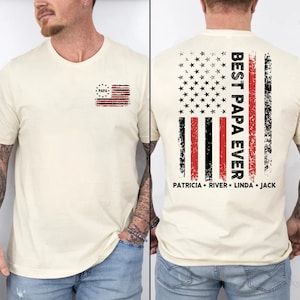 Puede incluir: Una camiseta de color crema con un dise&ntilde;o de bandera estadounidense desgastado en la espalda. La bandera tiene rayas rojas y negras con un dise&ntilde;o de estrella roja, blanca y azul en la esquina superior izquierda. El texto "BEST PAPA EVER" est&aacute; impreso verticalmente en el lado derecho de la bandera. Debajo de la bandera, el texto "PATRICIA * RIVER * LINDA * JACK" est&aacute; impreso. La parte delantera de la camiseta tiene un peque&ntilde;o dise&ntilde;o de bandera estadounidense desgastado con la palabra "PAPA" en el centro.