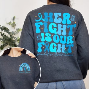 May include: A dark gray sweatshirt with a blue and teal design that says "Her Fight Is Our Fight" with butterflies and a ribbon. The text "Diabetes Awareness" is below the design. The front of the sweatshirt has a rainbow with a blue ribbon and the text "Team Marie".
