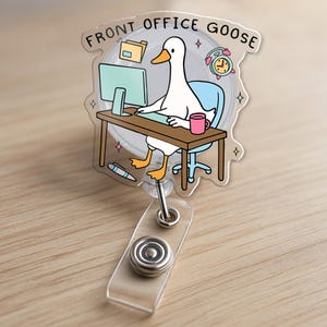 May include: A clear acrylic badge reel featuring a cartoon goose at a desk with the text "FRONT OFFICE GOOSE". The goose is depicted with a computer, coffee mug, and alarm clock. The reel has a retractable cord and a belt clip.