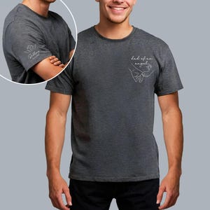 May include: Dark gray t-shirt with the text "dad of an angel" and a line drawing of two hands holding each other. The left sleeve has a small floral design and the name "Riley" in script. The shirt is made of a soft material.