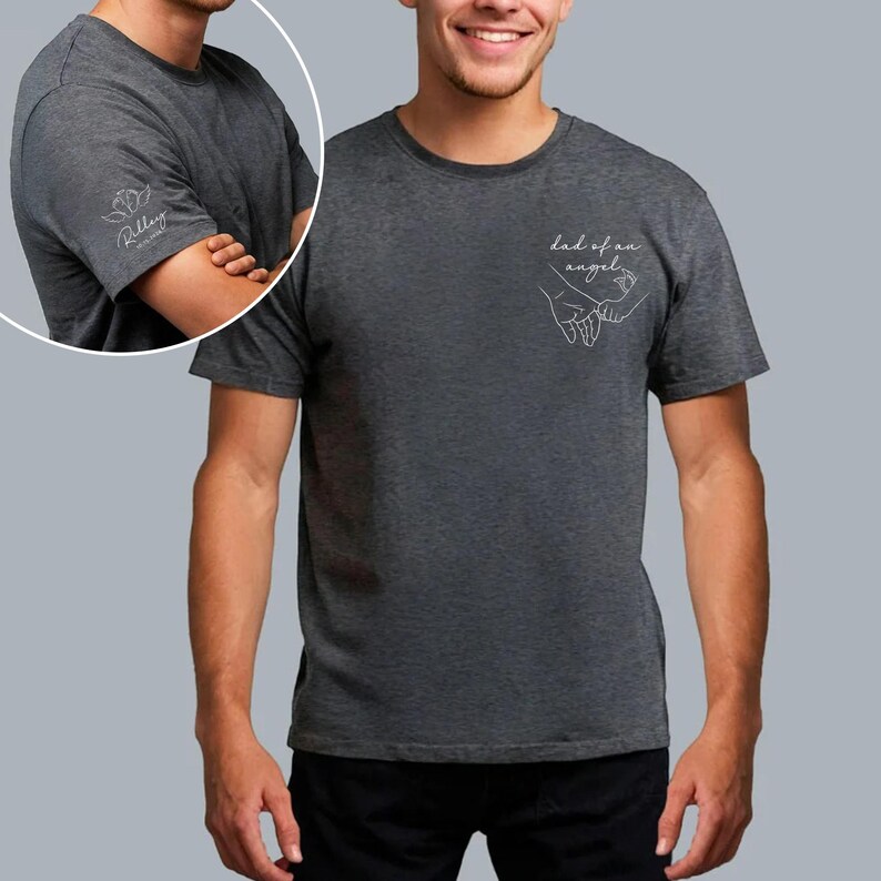 May include: Dark gray t-shirt with the text "dad of an angel" and a line drawing of two hands holding each other. The left sleeve has a small floral design and the name "Riley" in script. The shirt is made of a soft material.