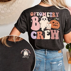 May include: A dark grey t-shirt with the text "Optometry Boo Crew" in a retro font. A ghost wearing glasses and holding an eye chart is next to a pumpkin. The back of the shirt has a smaller design with a ghost and the text "Spooky Optometrist Ashley".