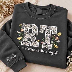 May include: A charcoal gray crewneck sweatshirt featuring the letters RT in a white lace pattern, surrounded by embroidered yellow and pink flowers. The text radiologic technologist is printed below, and the name Emily is embroidered on the sleeve.