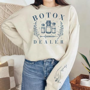 May include: Beige sweatshirt with the text "BOTOX DEALER" in navy blue, along with an illustration of vials and a syringe. The right sleeve features the name "Amelia" and a hand holding a syringe.