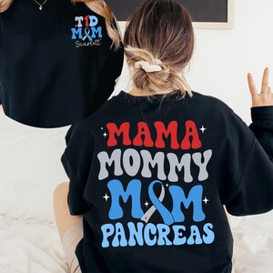 Pu&ograve; includere: Felpa nera con una grafica blu, rossa e grigia che dice "Mama Mommy M&M Pancreas". La grafica include un nastro blu con un centro bianco. La felpa ha anche una piccola grafica sul davanti che dice "T1D M&M Scarlett".