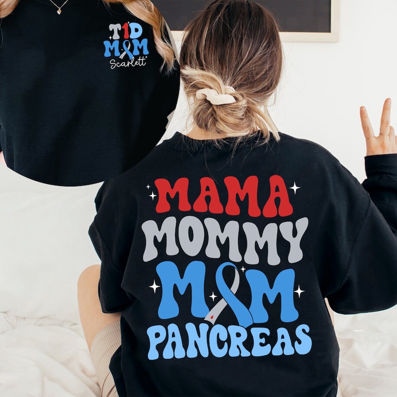 Pu&ograve; includere: Felpa nera con una grafica blu, rossa e grigia che dice "Mama Mommy M&M Pancreas". La grafica include un nastro blu con un centro bianco. La felpa ha anche una piccola grafica sul davanti che dice "T1D M&M Scarlett".