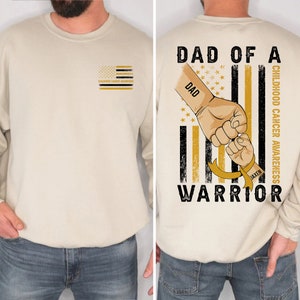 May include: A tan sweatshirt with a black and gold American flag design on the back. The flag has the words "Dad of a Warrior" and "Childhood Cancer Awareness" on it. The word "Dad" is on the left side of the flag and the word "Warrior" is on the bottom. The flag has a gold ribbon with the name "Jaxen" on it. The sweatshirt has a small black and gold American flag on the left chest with the words "Childhood Cancer Awareness" underneath it.