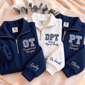 May include: Three quarter-zip sweatshirts in navy and white. The navy sweatshirts feature OT Occupational Therapist and PT Physical Therapist text with gingham and polka dot patterns. The white sweatshirt features DPT Doctor of Physical Therapy text with a floral pattern. Sleeves are embroidered with the names Emily, Dr. Sarah, and Olivia.