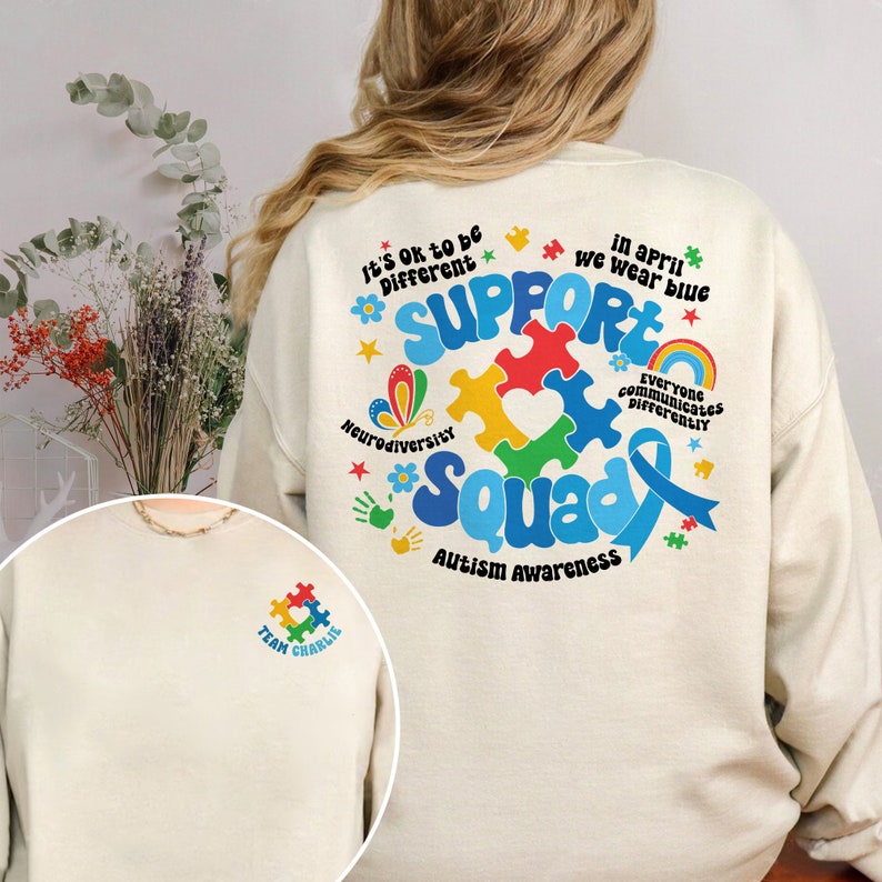 May include: A cream-colored sweatshirt with a blue ribbon, puzzle pieces, and the text "Support Squad" in rainbow colors. The text "It's Ok to be Different" and "Neurodiversity" are also included. The back of the sweatshirt has a small pocket with a puzzle piece and the text "Team Charlie".