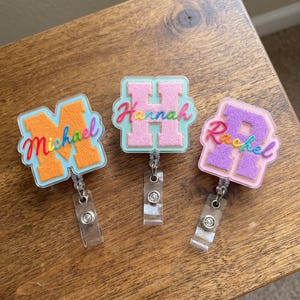 May include: Three badge reels featuring acrylic bases with textured varsity-style letters. The left reel has an orange M with the name Michael in rainbow script. The center reel has a pink H with the name Hannah in rainbow script. The right reel has a purple R with the name Rachel in rainbow script.