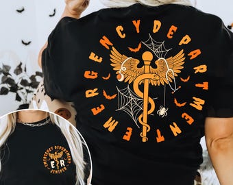 Op de afbeelding: Zwart T-shirt met een oranje en goudkleurig ontwerp met een medisch symbool, vleugels, vleermuizen en spinnenwebben. De woorden "Emergency Department" staan in een cirkel rond het ontwerp. Een kleinere versie van het ontwerp staat op de voorkant.