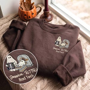 May include: A dark brown crewneck sweatshirt featuring an embroidered graphic on the left chest. The design shows two ghosts, one operating a CT scanner and another lying inside it, with the text "Jessica RT(R) Rad Tech" embroidered below.