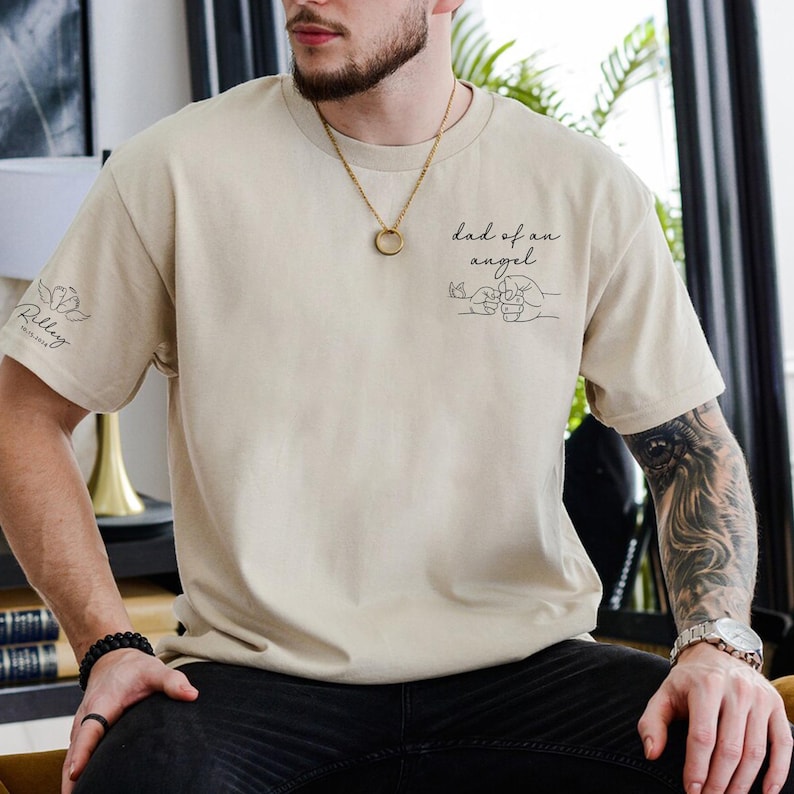 May include: Beige t-shirt with black text and line art. The front of the shirt says "dad of an angel" with two hands clasped together. The left sleeve has a floral design and the name "Riley" with a date.