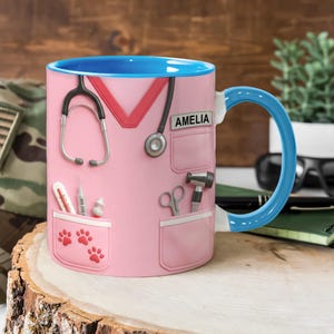 May include: Pink ceramic mug with a blue interior and handle. The mug is designed to resemble a medical uniform, complete with a stethoscope, medical tools, and the name "AMELIA". Features paw prints in the pocket.