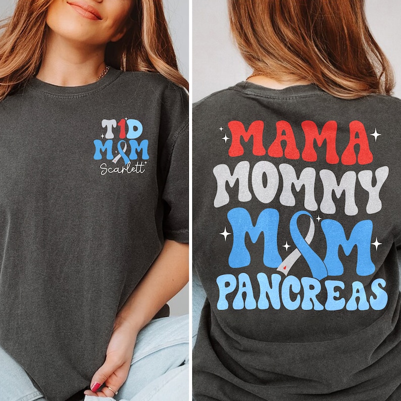 Pu&ograve; includere: Una maglietta grigio scuro con un nastro blu e il testo "T1D M&M Scarlett" sul davanti. La parte posteriore della maglietta ha il testo "Mama Mommy M&M Pancreas" in un font retr&ograve; con accenti rosso, bianco e blu.