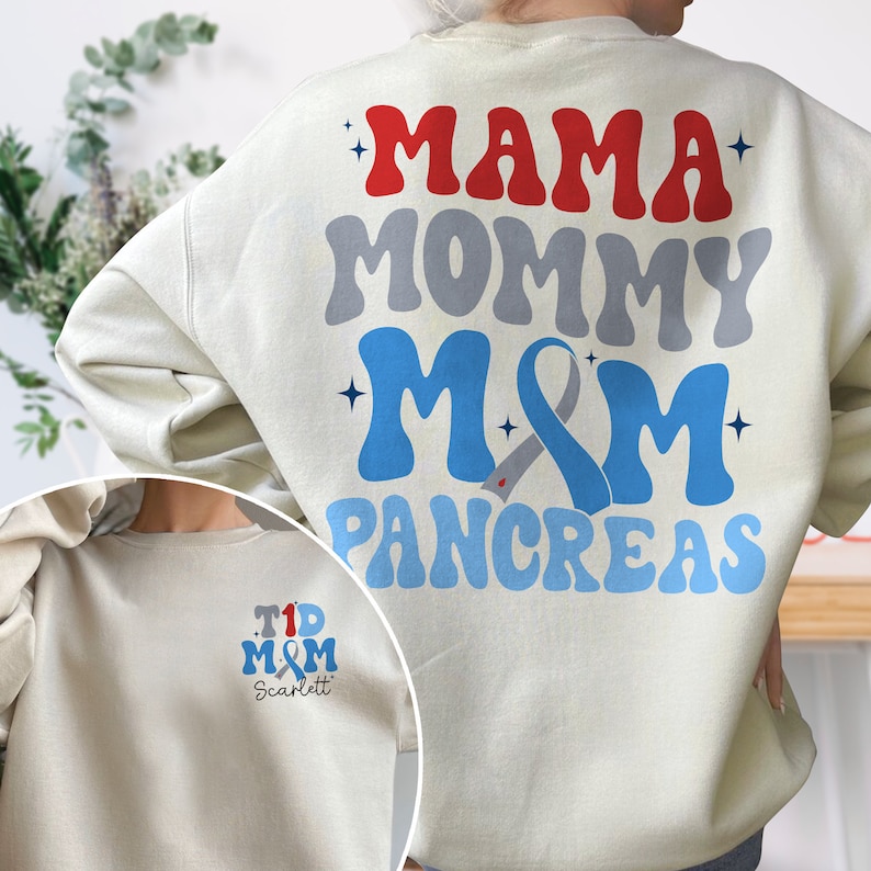 Pu&ograve; includere: Una felpa beige chiaro con il testo "Mama Mommy M&M Pancreas" stampato sul retro in rosso, grigio e blu. La parte anteriore della felpa ha un design pi&ugrave; piccolo con il testo "T1D M&M Scarlett" in blu e rosso.
