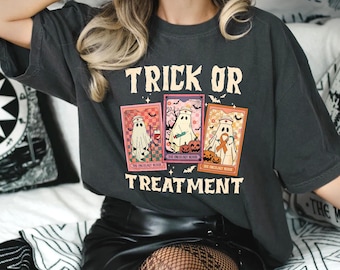 May include: Dark gray t-shirt with the words "TRICK OR TREATMENT" in a vintage font. The shirt features three tarot card-style illustrations of ghosts, pumpkins, and bats, with the text "THE ONCOLOGY NURSE".