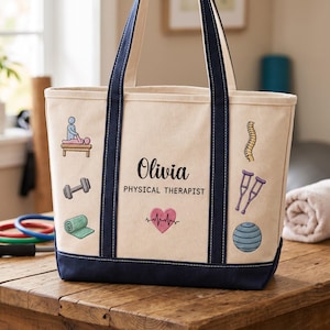 May include: A cream-colored canvas tote bag with navy blue handles and base. The bag features the text "Olivia" and "PHYSICAL THERAPIST" in black, surrounded by embroidered icons of a person on a table, a dumbbell, a mat, a spine, crutches, and a heart.